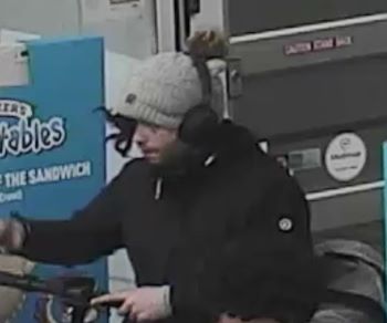 side view of male suspect; wearing a toque with headphones over top. His hair is flowing out of his toque to cover one eye. He is wearing a black coat.