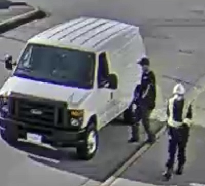 suspect one wearing white hard hat, a face mask, black shirt with a high visibility vest, black pants, black gloves, and black boots. Suspect two wearing black ball cap, black shirt, black pants, black boots, black gloves, a face mask, and glasses, standing beside the white Ford Econoline van.