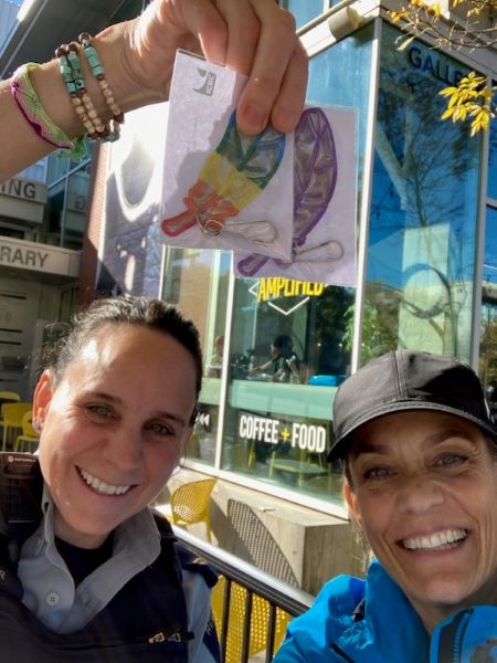 From left, Cpl. Dana Napier and ICBC Road Safety Coordinator, Ingrid Brakop hold up a rainbow coloured, feather shaped zipper pull reflector, while standing in front of a downtown coffee shop with yellow writing in the window. 