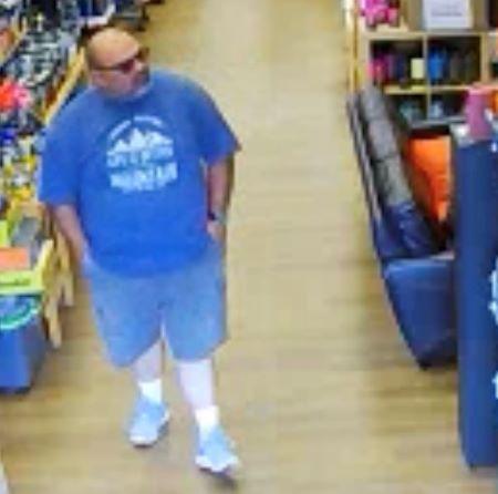 A middle aged Caucasian man, heavy set, bald on top with grey and black hair on the sides, as well as in his moustache and beard, walks around a retail store with his hands in his pockets. The man is wearing dark sunglasses, a blue t-shirt, and shorts. 