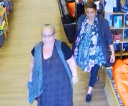 On the left, a Caucasian woman with grey hair, glasses, a dark blue or purple dress, and a blue vest walks around a store. A female with a media complexion, dark hair, long blue and pink floral shirt, and dark capris and shoes, walks behind her. 