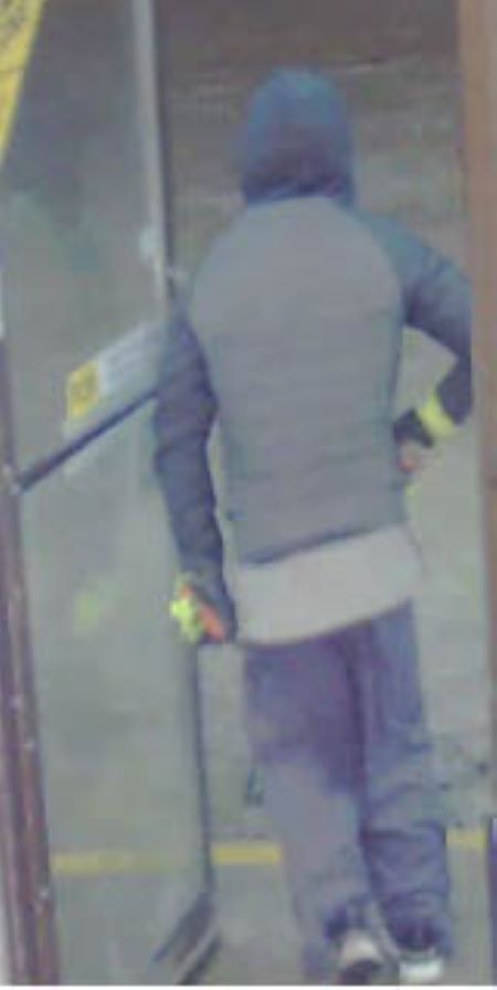 Suspect vu de dos : un homme de corpulence moyenne sortant par une porte vitrée et portant un manteau foncé, un pantalon et des gants ayant du jaune fluorescent.