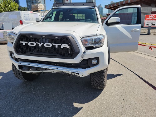 Front of a white Toyota Tacoma with frond end damage and driver’s door open. The word TOYOTA is written in white on the black front grill of the truck.