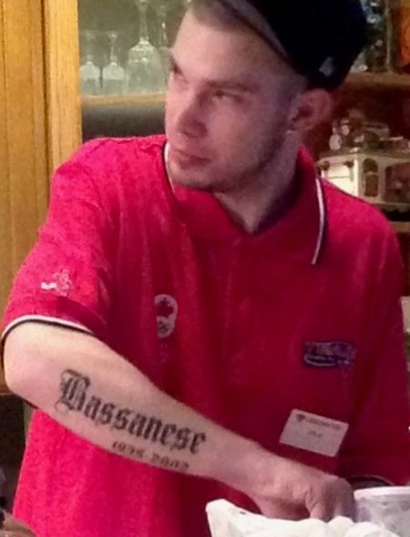 Kyle Ferg, coiffé d’une casquette noir et vêtu d’un haut rouge à manches courtes. On voit le tatouage « Bassanese » sur son avant-bras droit. 