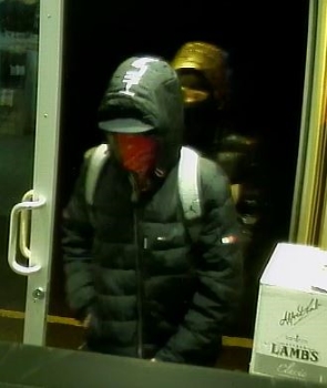 Le premier suspect entrant dans le magasin. Il porte un manteau noir dont le capuchon est relevé et transporte un sac à dos muni de bretelles grises. 