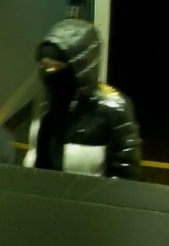 Le deuxième suspect entrant dans le magasin. Il porte un manteau noir et blanc brillant dont le capuchon est relevé. 