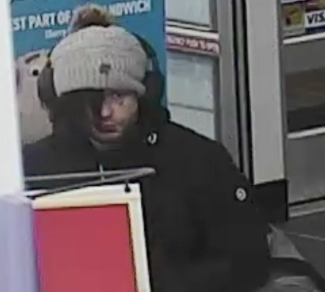 male suspect facing toward the camera; wearing a toque with headphones over top. His hair is flowing out of his toque to cover one eye. He is wearing a black coat. 