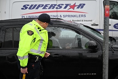 A man sitting in his vehicle at a red light using his cell phone. A police officer wearing a ball cap and yellow jacket observe the man using his cell phone. The vehicle is stopped in traffic and the police officer is on the passenger side of the vehicle. 